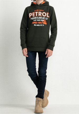 Grøn hættetrøje med "PETROL"-logo i hvid og orange, kombineret med mørke jeans og lysebrune sko. Hættetrøjen har snøre og et afslappet design.