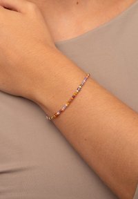 Farbiger Perlenarmband mit Mustern aus kleinen runden und sechseckigen Perlen in Lila, Orange, Grün und Gold, getragen am Handgelenk.