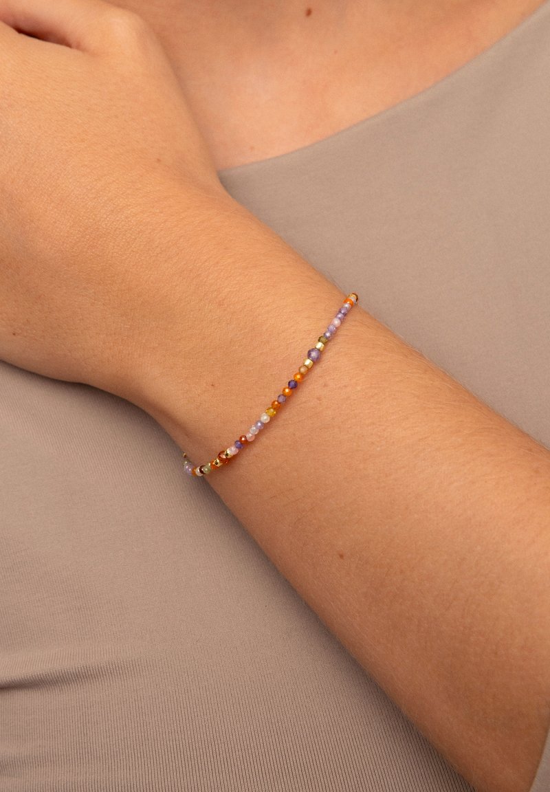 Farbiger Perlenarmband mit Mustern aus kleinen runden und sechseckigen Perlen in Lila, Orange, Grün und Gold, getragen am Handgelenk.