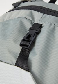 Grå tekstilryggsekk-seksjon med en strukturert finish, som har en svart plastspenne og webbed stropp, som fremhever funksjonelle designelementer.