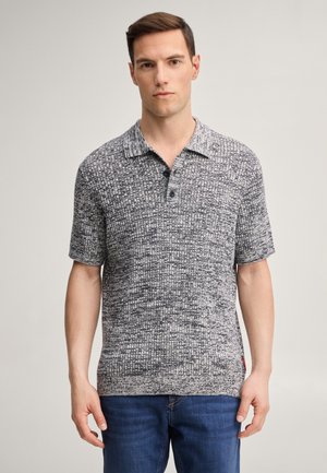 Mężczyzna z krótkimi brązowymi włosami nosi teksturowaną szarą dzianinową koszulkę polo z krótkim rękawem i niebieskie dżinsy, stoi na tle jednolitego jasnego tła.