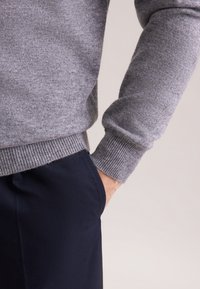 Grauer Pullover mit geripptem Saum und Bündchen, kombiniert mit dunkelblauen Hosen. Der Stoff wirkt weich mit einer subtilen Textur. Die Hand ist in die Tasche gesteckt.