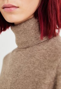Bruine geribde coltrui met een zachte textuur, nauwsluitend ontwerp en hoge hals, gecombineerd met diep rood haar.