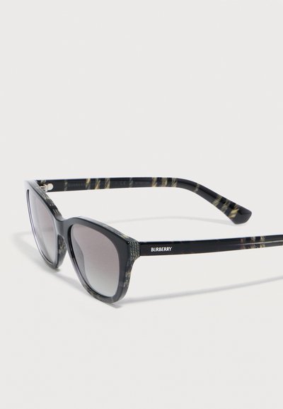 Lunettes de soleil Burberry noires à monture œil-de-chat avec verres dégradés foncés et motif discret à carreaux sur la monture et les branches, inclinées sur une surface blanche.
