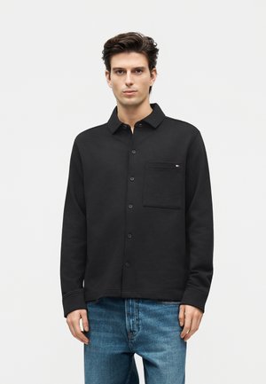 Άνδρας με μαύρο πουκάμισο με κουμπιά και τσέπη στο στήθος και μπλε τζιν, στέκεται μπροστά σε μονόχρωμο ανοιχτό φόντο.