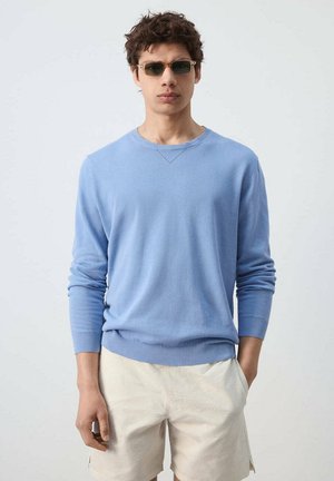 Joven con gafas de sol rectangulares, suéter azul claro de manga larga y pantalones cortos beige, de pie con una mano en el bolsillo.