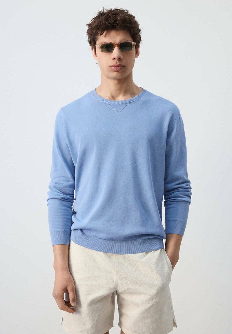 Jeune homme portant des lunettes de soleil rectangulaires, un pull bleu clair à manches longues et un short beige, debout avec une main dans la poche.