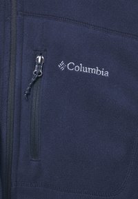 Navy zilā fleeces jaka ar rāvējslēdzēja kabatu, kontrastējošiem šuvēm un baltu Columbia logotipu uz krūtīm. Mīksta tekstūra un viegls dizains.