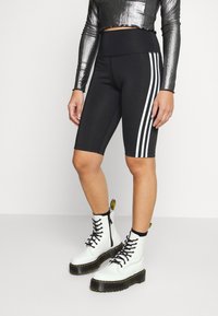 Shorts de cyclisme noirs avec trois bandes verticales blanches sur le côté ; associés à un haut métallisé à manches longues et des bottes blanches à lacets.