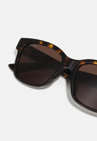 Ochelari de soare cu rame pătrate voluminoase, din material tartan, cu lentile maro închis și o marcaj discret pe marginea superioară. Suprafață netedă, design modern.