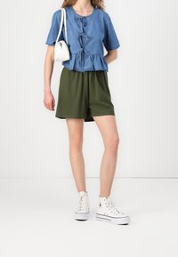 Top in denim a peplo con nodo frontale; pantaloni corti verdi casual; sneakers bianche alta. Tessuto leggero, design casual, colori a contrasto.