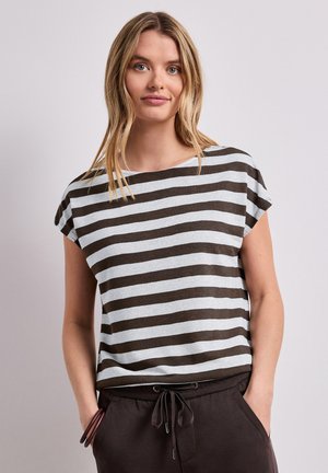 Młoda kobieta o długich blond włosach, ubrana w czarno-biały, poziomo paskowany krótki rękaw oraz ciemne spodnie ze sznurkiem, z rękami w kieszeniach.