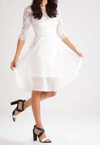 Vit klänning med spetsärmar i tre fjärdedelar, flytande kjol och figursydd bodice, tillsammans med svarta och vita ankelsnäckor med hög klack.