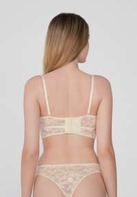Krajkové podprsenka v světlé žluté barvě s nastavitelnými ramínky, zapínáním na háček a očko a složitým květinovým vzorem, párovaná s našitým krajkovým tanga kalhotkami.
