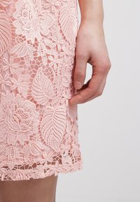 Main posée à côté d'une jupe en dentelle rose avec une broderie détaillée de fleurs et de feuilles sur un fond clair.