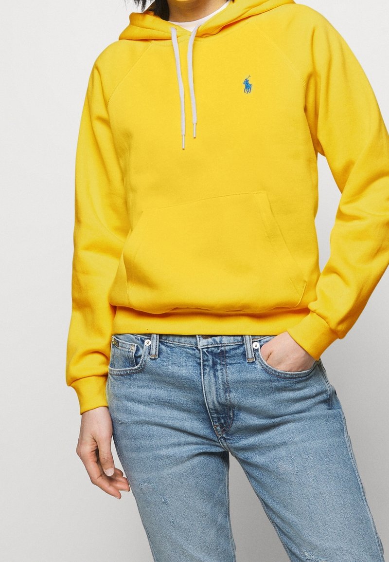 Gul hoodie med framficka, snörning och liten blå logotyp. I kombination med ljusblå slitna jeans.