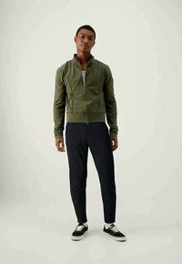Giacca verde con zip e cuciture testurizzate, abbinata a pantaloni blu scuro e sneakers nere. La giacca ha un colletto alto e un design aderente.