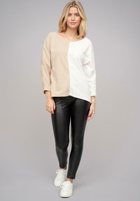 Beige en witte gesplitste lange mouwtop van zachte gebreide stof; gecombineerd met glanzende zwarte leggings. Model draagt witte sneakers.