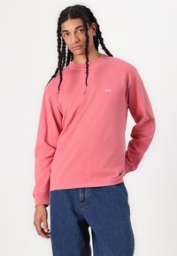 Long-sleeve, crew-neck sweatshirt in faded pink cotton with a white "obey" logo on the chest, worn with blue denim pants.