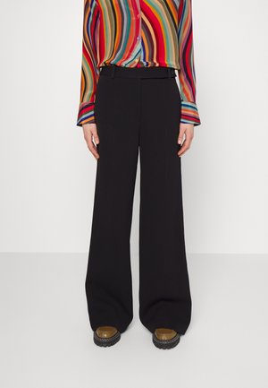 Persoon draagt zwarte broek met hoge taille en wijde pijpen, een multicolor gestreept shirt met lange mouwen, en bruine platformschoenen met zwarte zolen.