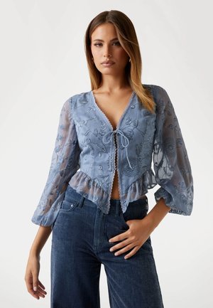 Light blue blouse with embroidered floral pattern, sheer long sleeves, tie detail at the front, and a ruffled hem. Paired with dark denim jeans.
