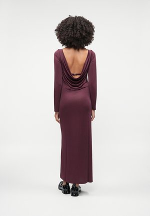 Femme aux cheveux bouclés portant une robe violette longue manches longues, longue jusqu'au sol, avec un dos ouvert et des chaussures noires brillantes, debout de dos.