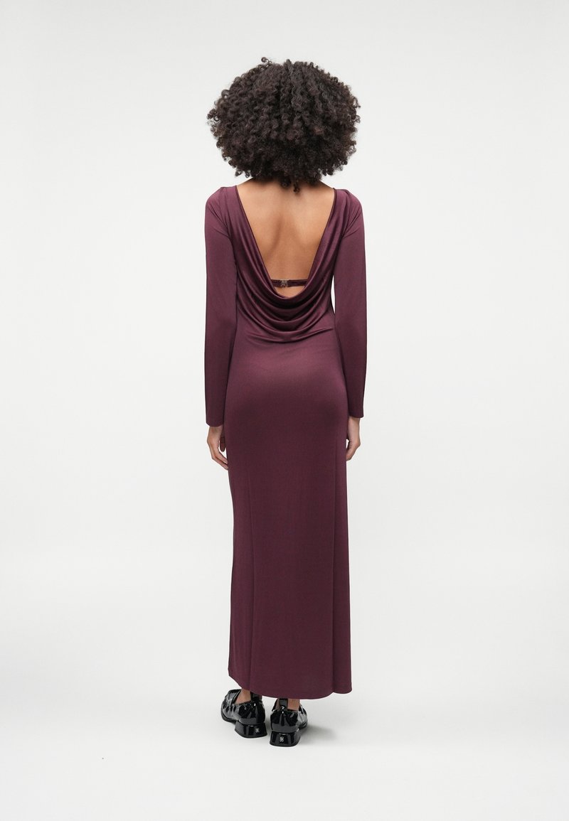 Femme aux cheveux bouclés portant une robe violette longue manches longues, longue jusqu'au sol, avec un dos ouvert et des chaussures noires brillantes, debout de dos.