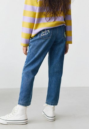 Denim bikses ar brīvu piegriezumu, zilā krāsā, ar izšūtu "Smiley World" uz aizmugures kabatas. Saskaņotas ar baltām augstām kedām.