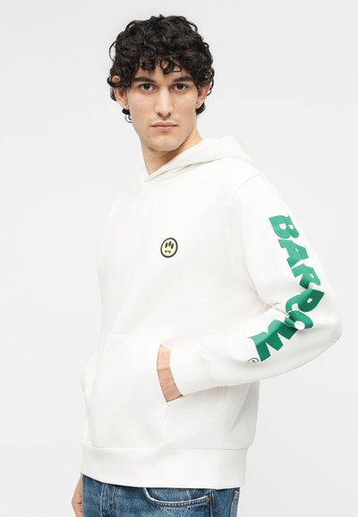 Νεαρός άνδρας με σγουρά σκοτεινά μαλλιά που φοράει λευκό φούτερ με κουκούλα με ένα μικρό κίτρινο χαμογελαστό πρόσωπο στο στήθος και πράσινο κείμενο στο μανίκι.