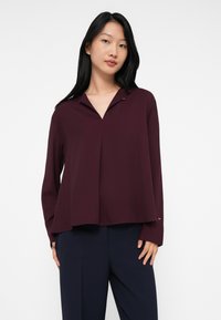 Woman with long black hair wearing a loose, long-sleeve dark purple blouse and navy blue pants against a plain white background.