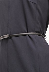 Czarny skórzany pasek z metalową prostokątną klamrą, noszony na ciemnym materiałowym ubraniu. Pasek ma gładką teksturę i smukły profil.