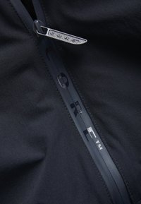 Veste imperméable noire avec une fermeture éclair lisse, un ruban de marque avec le texte "CORE" et un tissu texturé. La tirette de la fermeture éclair a un design distinct.