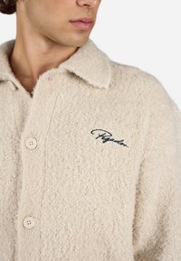 Beige, fuzzy cardigan med krave, der har to store knapper og et broderet logo i sort på brystet. Blød tekstur.