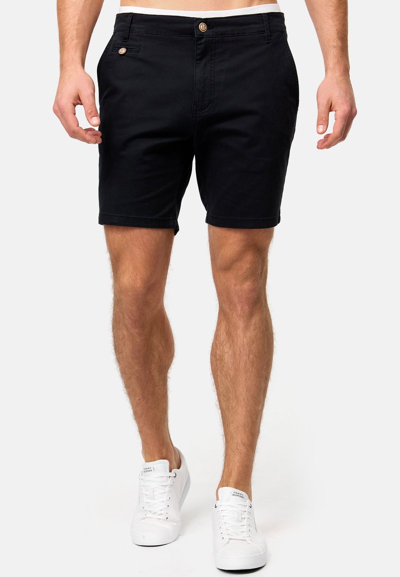 Shorts en coton noir avec fermeture à bouton, poches latérales et coupe droite. Portés avec des baskets blanches.