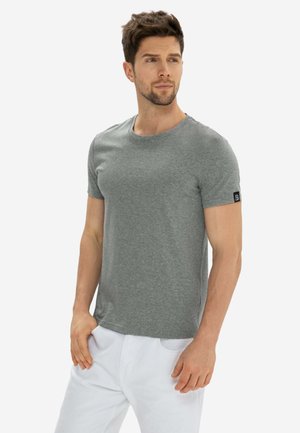 Uomo con capelli corti castani che indossa una maglietta grigia a maniche corte e pantaloni bianchi, guarda leggermente verso destra.