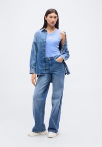 Jeune femme portant un débardeur bleu clair, une chemise en jean oversize, un jean à jambes larges et des baskets blanches, debout avec une main dans la poche.
