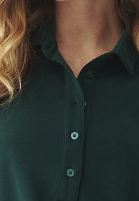 Ciemnozielona koszula z kołnierzykiem, wykonana z miękkiego materiału, z zapięciem na guziki. Posiada widoczne szwy i cztery okrągłe guziki na listwie.