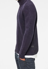 Stickad polotröja i mörklila, med upphöjda horisontella ränder och en figurnära passform. Märke på den nedre sömmen.