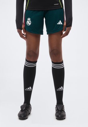 Dolna część ciała osoby ubranej w ciemnozielone spodenki piłkarskie Real Madrid, czarne, adidasowe skarpety do kolan i czarne korki na białym tle.
