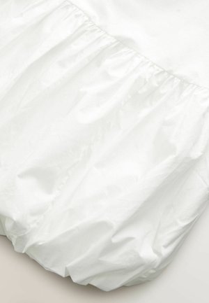 Balta suknelė, kurią puošia raukšlėtas volanas apatinėje dalyje, pagaminta iš lengvos, lygaus audinio su šiek tiek blizgiu paviršiumi.