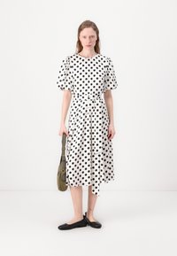 Vestido branco com grandes bolinhas pretas, mangas curtas em balão, cintura com cinto e comprimento até ao joelho. Combinado com sapatos pretos e uma mala verde-oliva.