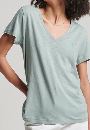 Kobieta nosząca jasnooliwiany krótki rękaw T-shirt z dekoltem w serek i białe spodnie, z ręką spoczywającą na biodrze.