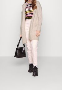 Donna con cardigan beige, top multicolore a righe, pantaloni rosa chiaro, sneakers nere, che tiene una borsa nera con texture su uno sfondo semplice.