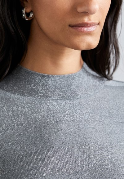 Tejido texturizado plateado con un cuello alto, diseño ajustado y un brillo sutil. Presenta patrones geométricos y pendientes minimalistas plateados.