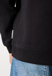 Nahaufnahme einer Person, die einen schwarzen Kapuzenpullover mit einem kleinen weißen Etikett und hellblauen Jeans trägt.