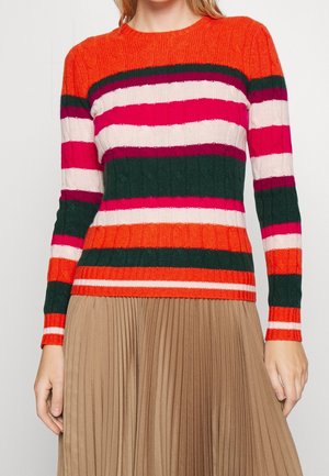 Vrouw draagt een kabelgebreide trui met lange mouwen met horizontale oranje, groene, roze en witte strepen, gecombineerd met een beige geplooide rok.
