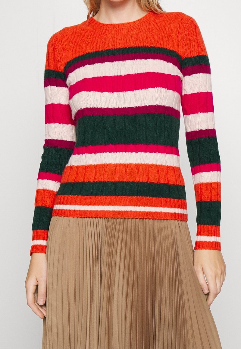 Femme portant un pull en maille torsadée à manches longues avec des rayures horizontales orange, vert, rose et blanc, associé à une jupe plissée beige.