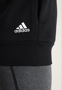 Schwarzer Kapuzenpullover mit geripptem Saum und weißem Adidas-Logo. Kombiniert mit grauen, strukturierten Leggings mit sichtbarer Naht.