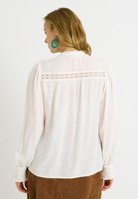 Blusa blanca de manga larga con tela texturizada, hombros fruncidos y detalles de encaje en la espalda. Puños elásticos que realzan el diseño.