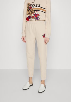 Pantalon de jogging beige avec broderies florales et tigre près de la poche, porté avec un top assorti et des chaussures blanches à enfiler.